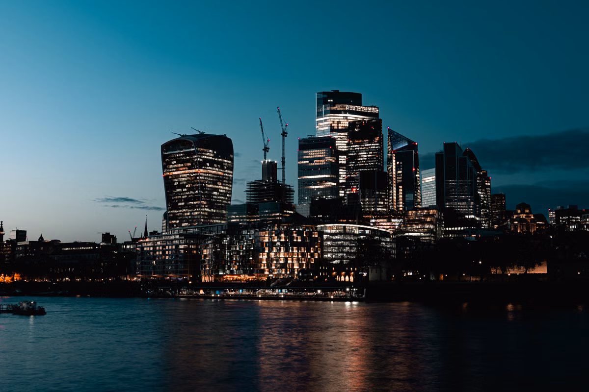 The City of London's office towers lit up at dusk, seen across the River Thames, with cranes above the skyline.