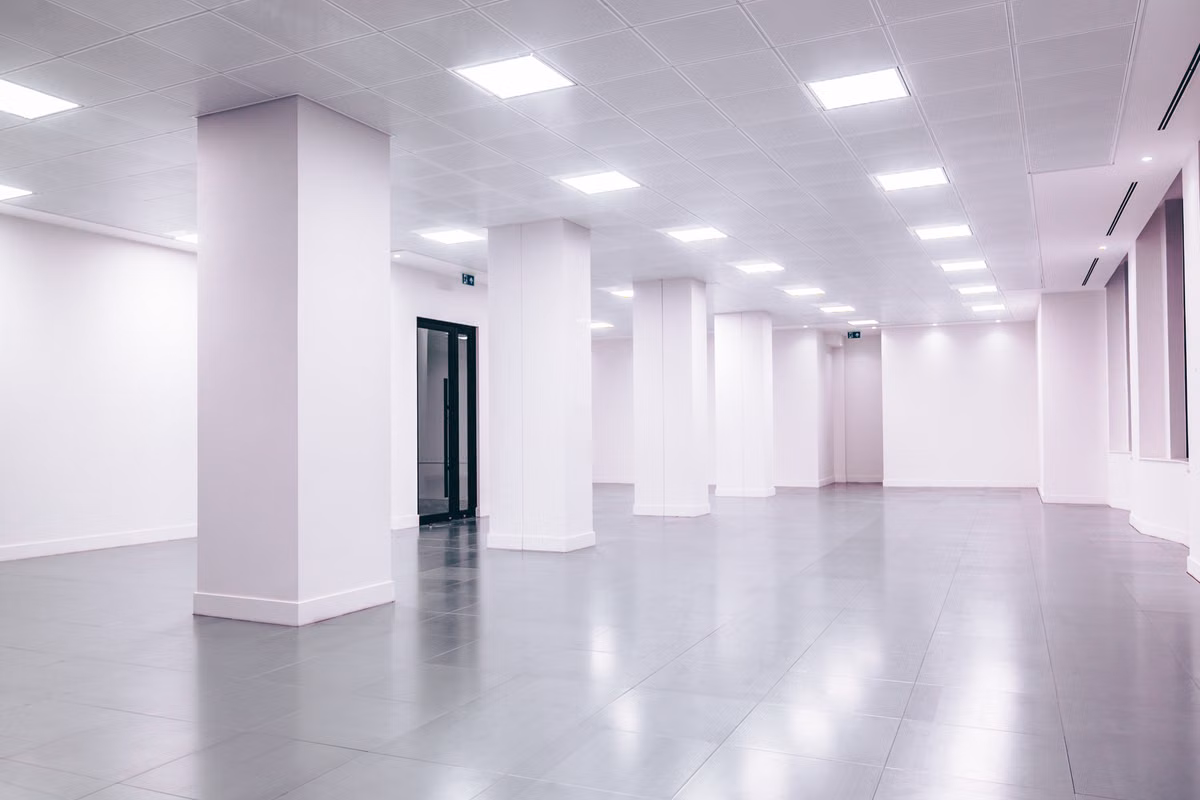 An empty open-plan interior with white columns, a grey tiled floor and a suspended grid ceiling fitted with recessed square light panels.