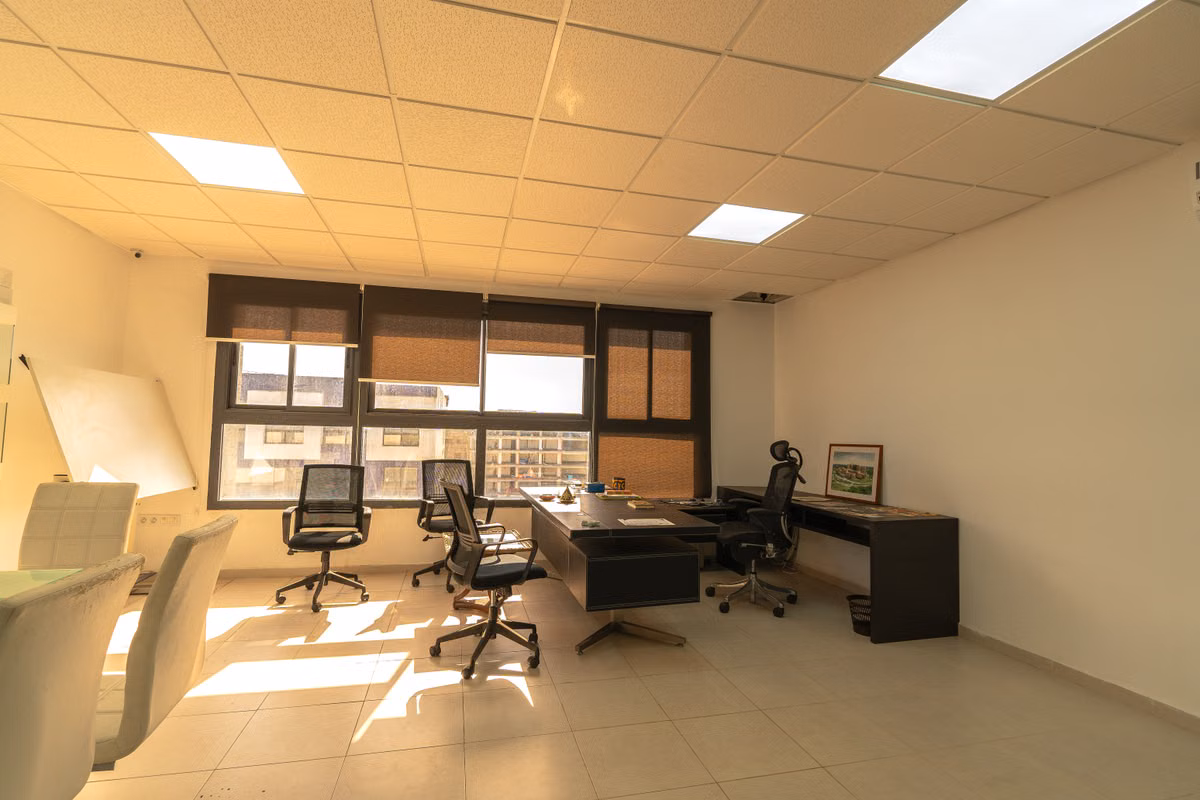 An empty office with a suspended tiled ceiling, recessed panel lights, dark desks, office chairs and window blinds.