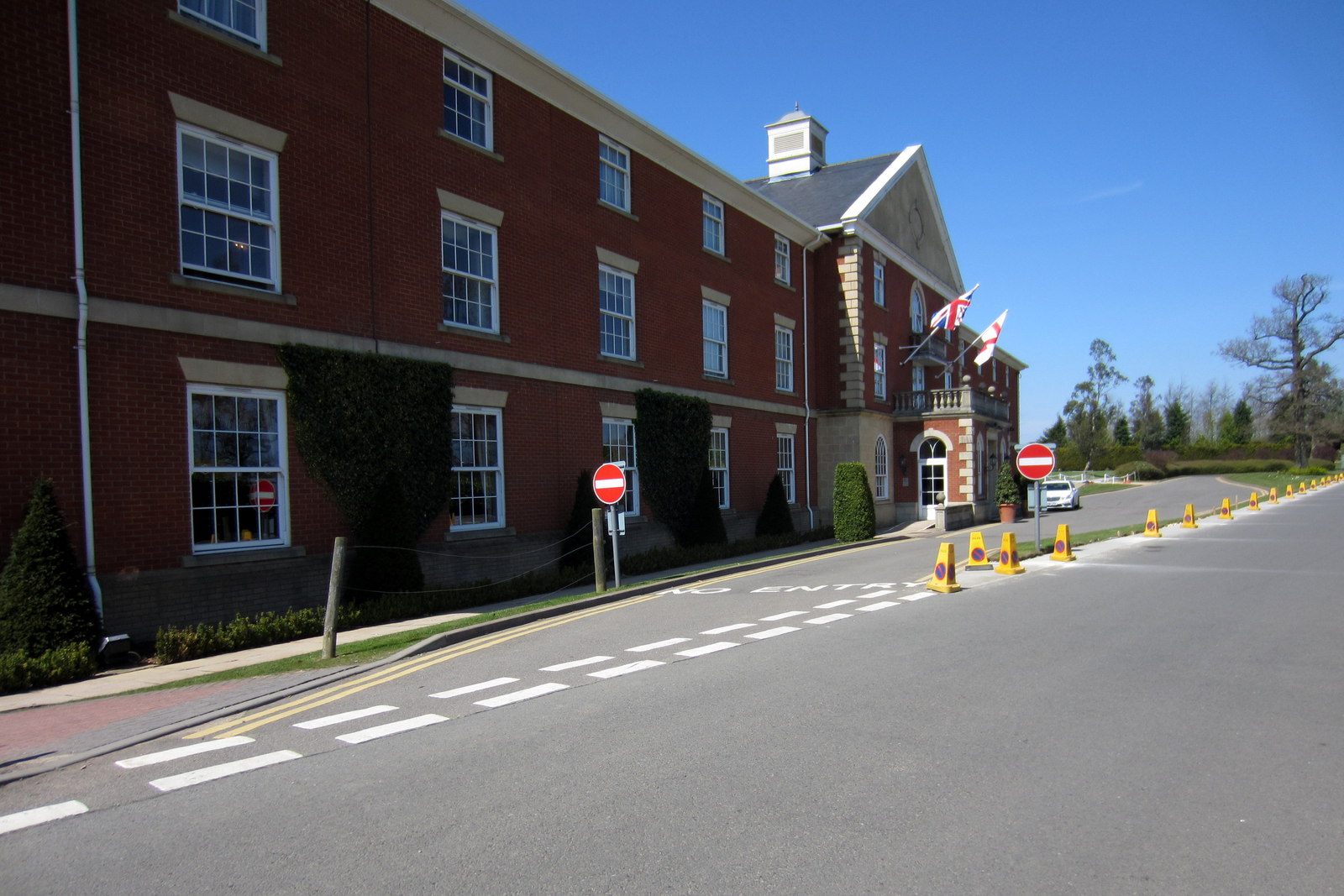 A three-storey red-brick hotel building with sash windows and Union and St George flags over its entrance, beside an access road lined with traffic cones.