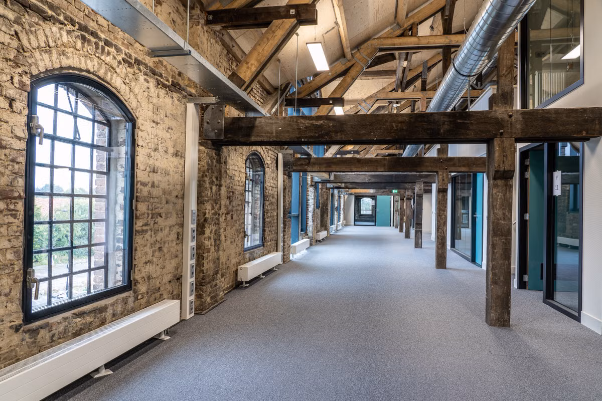 An empty open-plan office floor inside a converted brick building, with exposed brick walls, arched windows, timber roof trusses, overhead ducting and glass-fronted offices.