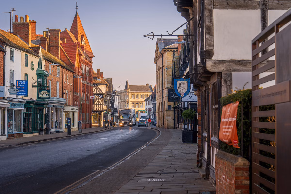 A quiet English town high street in early morning light, with historic brick and timber-framed buildings and ground-floor shopfronts on both sides of the road.