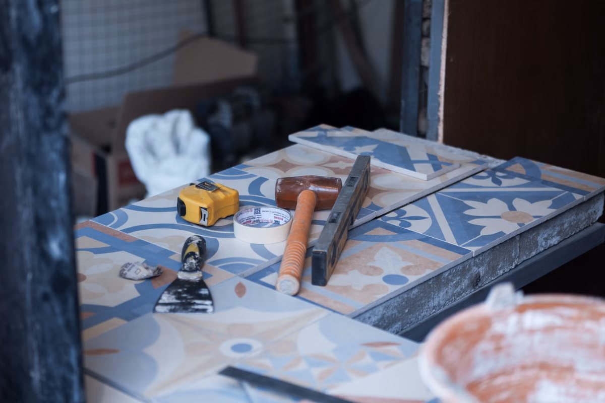 Tiling tools including a tape measure, rubber mallet, spirit level, masking tape and a scraper laid on patterned blue and cream floor tiles.